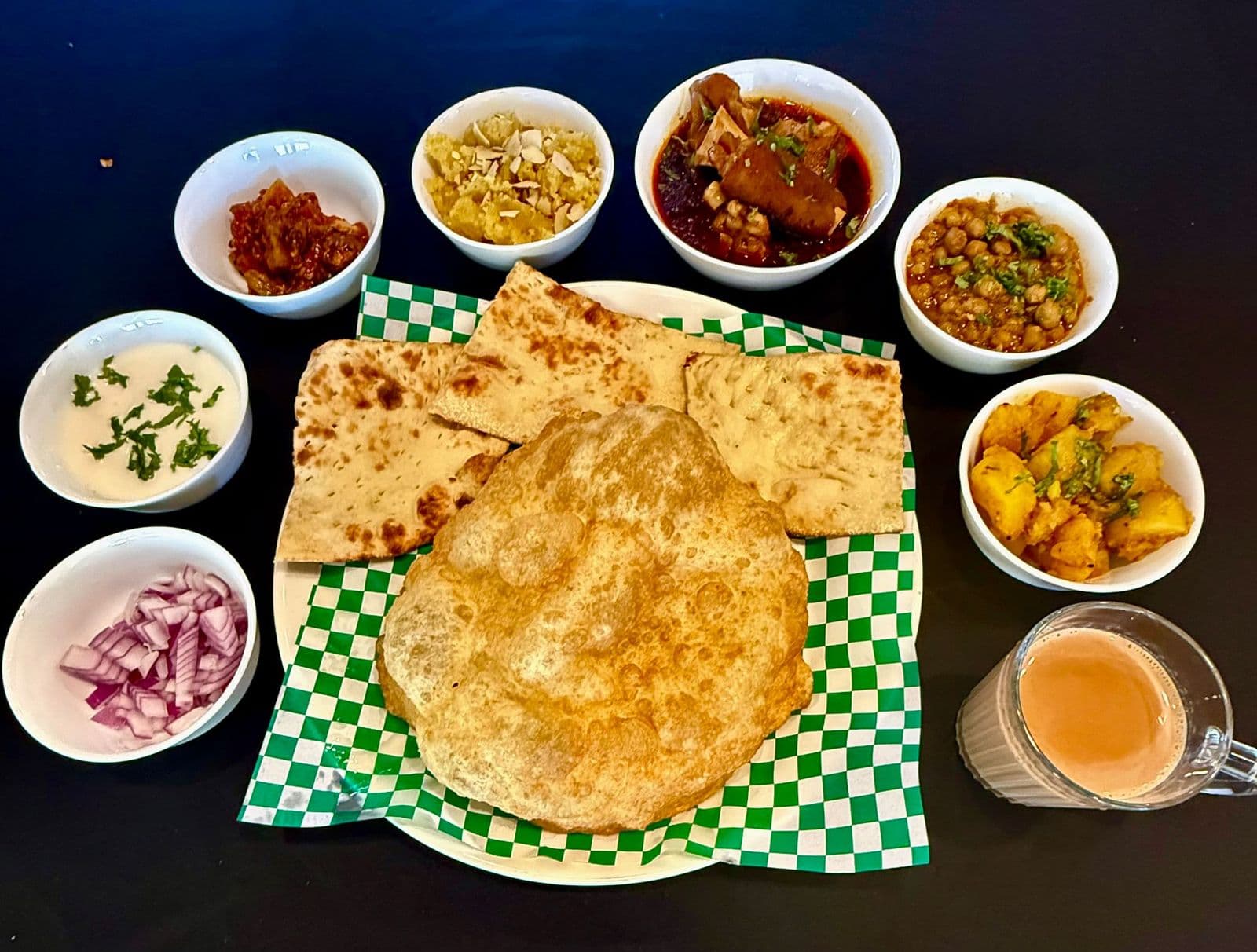 The Weekend Halwa Puri Brunch spread: puffed puri and naan with paya, chana, aloo, halwa, pickle, yogurt, onions and a glass of karak chai