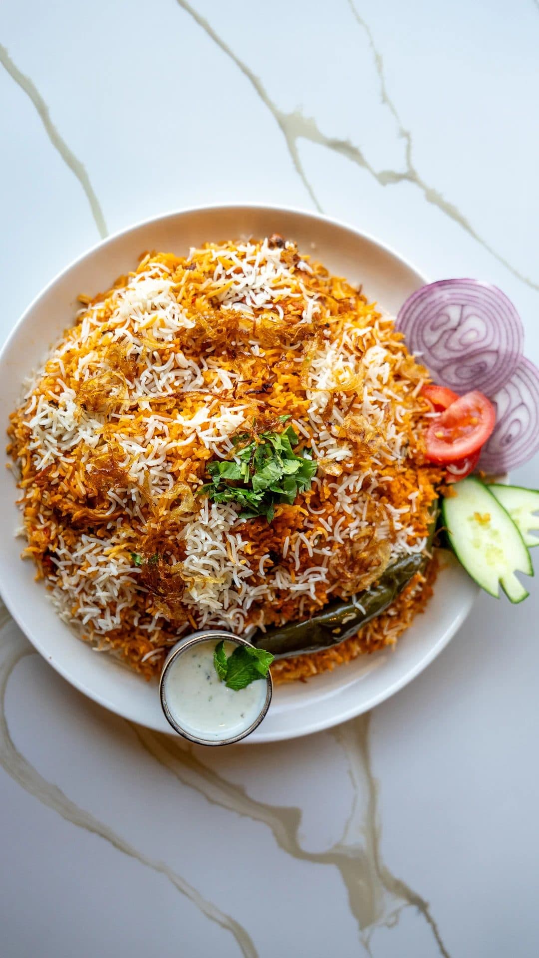 Plate of spicy Karachi biryani rice topped with fried onions, served with raita, green chili, onion and cucumber